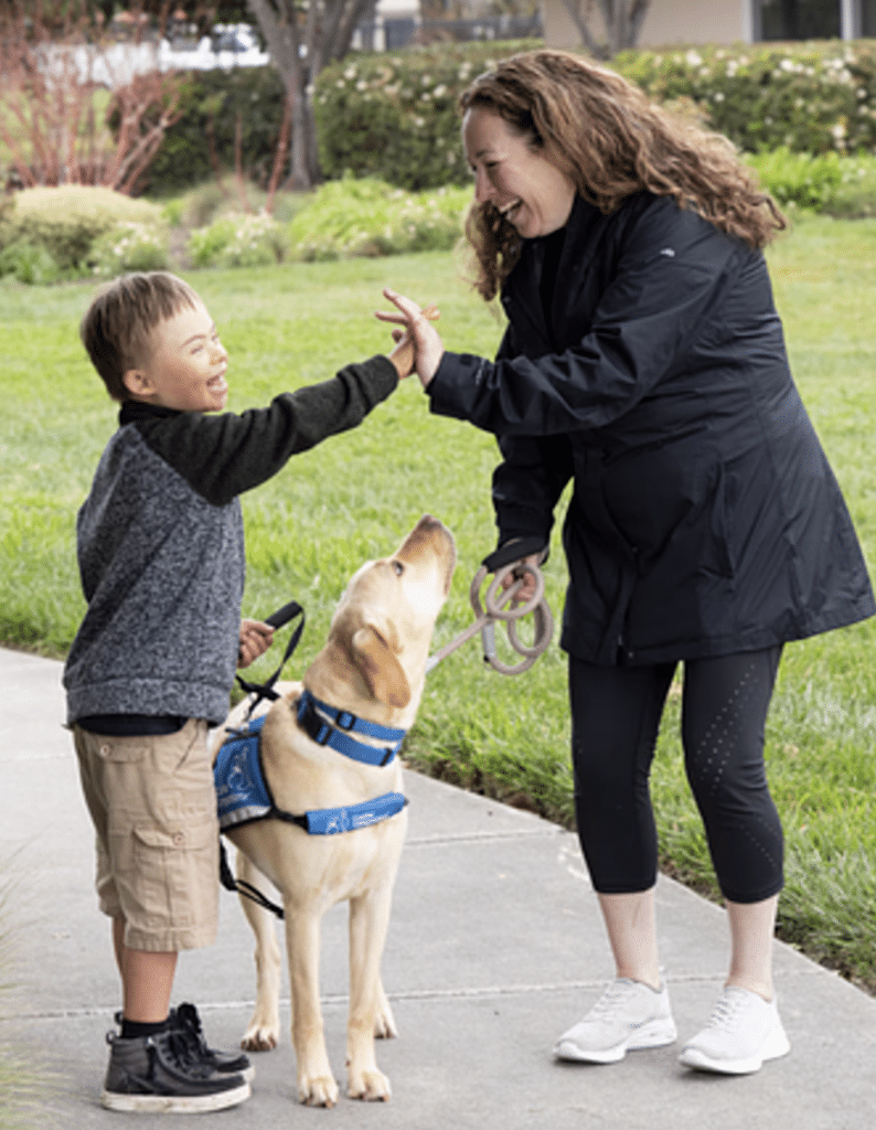 A woman and child share a high-five outdoors with a yellow Lab wearing a blue assistance dog vest.